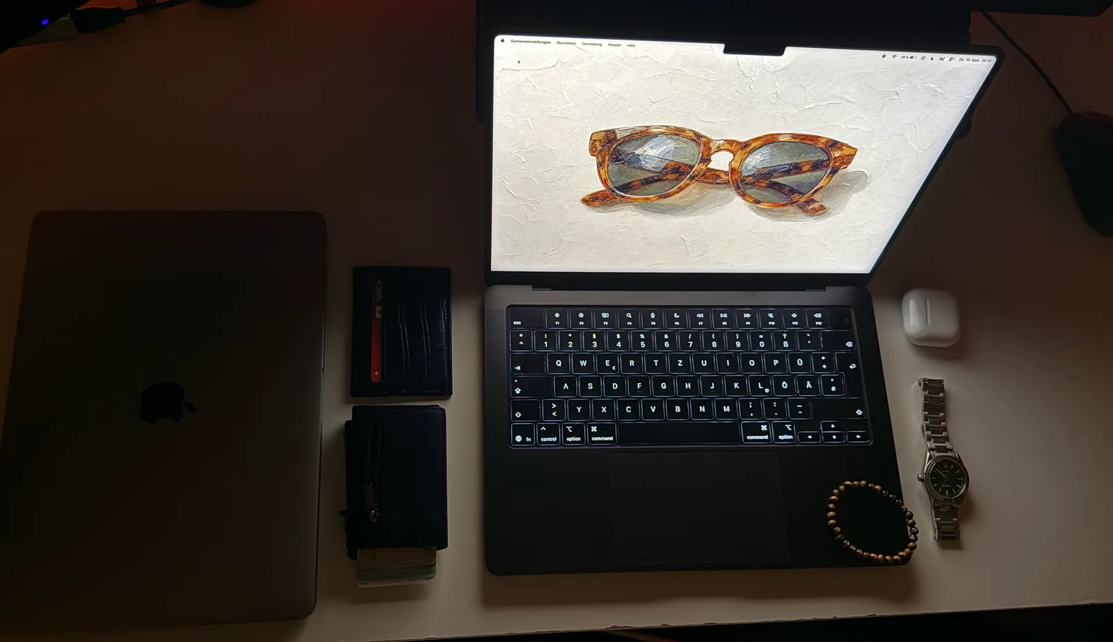 Schreibtisch von oben: aufgeklapptes MacBook mit dem Sonnenbrillen-Motiv, daneben ein zugeklapptes Notebook, Kartenetuis, Uhr und AirPods.